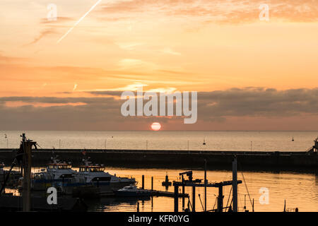 Sun salito sopra l'orizzonte oltre la Manica. Il sole è una sfera rossa appena circa per agganciare un strato di nube spessa sopra. Orange sky. Ramsgate Harbour in primo piano. Foto Stock