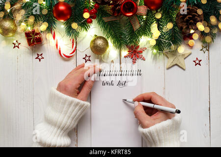 Donna con le mani in mano la scrittura su un blocco note, Natale la lista dei desideri su bianco tavolo in legno con ornamens festosa e luci.spazio copia,su testa piana vista lay Foto Stock