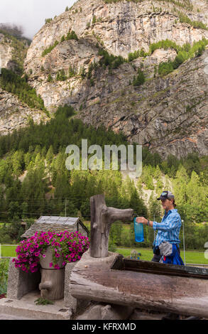 Herbriggen, Svizzera - escursionista si riempie di acqua in bottiglia in acqua pubblica fontana con scogliera sul retro. Foto Stock