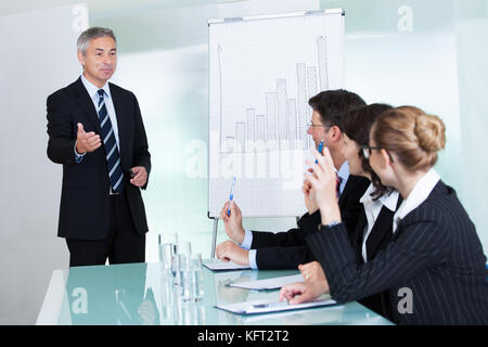 Manager o senior business executive in piedi di fronte a un grafico dando una presentazione al personale o ai colleghi seduti a un tavolo Foto Stock