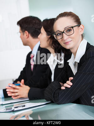 Bella giovane imprenditrice con un sorriso affascinante che indossano occhiali guardando la telecamera mentre in occasione di un incontro con i colleghi Foto Stock