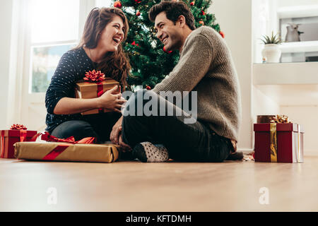 Felice coppia giovane con confezioni regalo seduto vicino a un albero di Natale. Giovane uomo e donna per celebrare il Natale a casa. Foto Stock