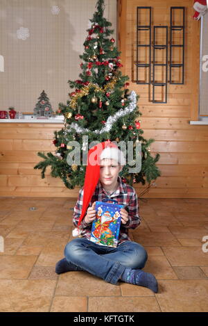 Il ragazzo si siede sul pavimento e ritiene che il dono di un albero di natale. Foto Stock