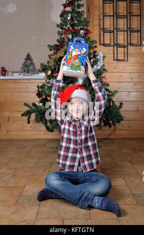 Il ragazzo si siede sul pavimento e tiene nelle sue mani tese un regalo a un albero di natale. Foto Stock