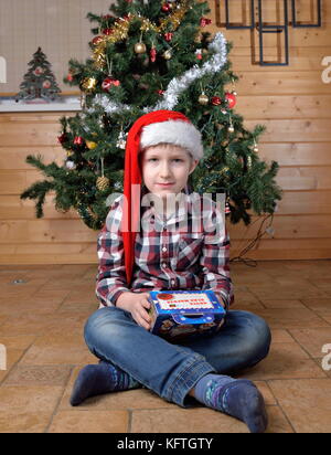 Il ragazzo si siede sul pavimento e ritiene che il dono di un albero di natale. Foto Stock