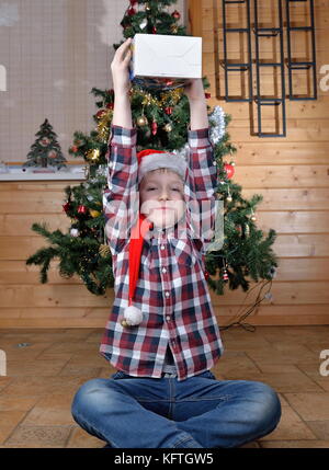 Il ragazzo si siede sul pavimento e tiene nelle sue mani tese un regalo a un albero di natale. Foto Stock