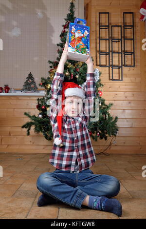 Il ragazzo si siede sul pavimento e tiene nelle sue mani tese un regalo a un albero di natale. Foto Stock