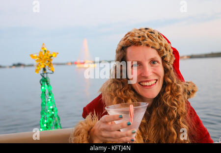 Signora giovane in una festosa vacanza santa hat detiene un candy cane martini. decorate albero e barca in background. Foto Stock