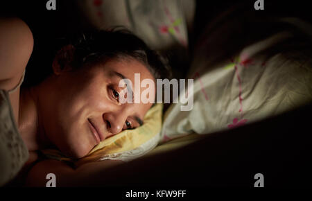 Donna sorridente con tablet nel letto prima di dormire. luce brillante da digtal compressa sulla faccia ragazza nel letto Foto Stock