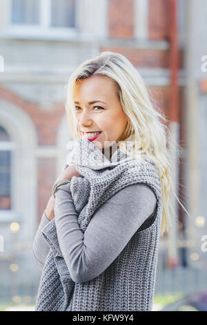 Giovane donna bionda con cardigan grigio che cammina per la vecchia strada della città. Moda casual, look elegante Foto Stock