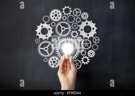 Mano che tiene una lampadina anteriore ingranaggi su disegno di Blackboard Foto Stock