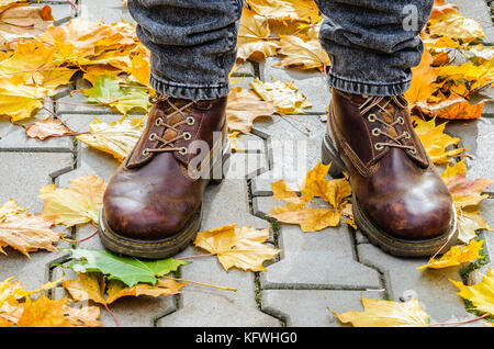 Gambe in pelle marrone stivali tra caduto foglie di acero in grigio di una pavimentazione in cemento. a piedi nel parco d'autunno. Foto Stock