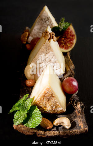 Pezzi di formaggio ammuffito con uva, menta, fichi, mandorle, anacardi sulla corteccia su sfondo nero. Alimentare la composizione per lo styling con formaggio essiccato. Foto Stock