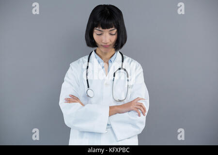 Riflessivo medico in piedi con le braccia incrociate contro uno sfondo grigio Foto Stock