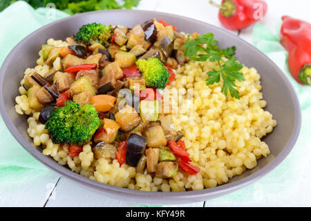 Vegano caldo con insalata di cuscus e verdure fritte (melanzane, zucchine, cipolla, peperone, broccoli) in un recipiente di ceramica su un bianco sullo sfondo di legno Foto Stock