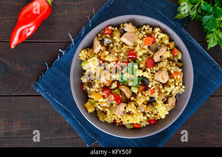 Luce dieta sana insalata con cuscus, verdure (zucchine, melanzane, carote, peperoni, cipolle), pezzetti di pollo al buio su un sfondo di legno Foto Stock