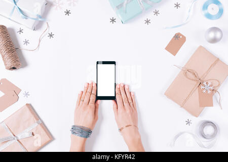 Mani femminili in possesso di un telefono cellulare fra le decorazioni di Natale e fiocchi di neve su un tavolo bianco vista superiore Foto Stock