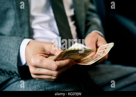 Un giovane uomo caucasico in un elegante abito grigio conta euro bills seduto nel sedile posteriore di una macchina Foto Stock