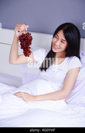 Donna con uva rossa sul letto in camera da letto Foto Stock