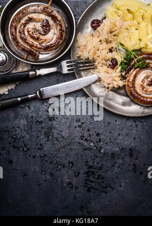 Salsicce fritte con patate e cavolo sott'aceto servita su dark tavolo rustico sfondo con posate, vista dall'alto Foto Stock