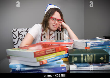 Giovani annoiati e stanchi femmina ritratto infermiere bianco da indossare scrubs, tappo con assistenza infermieristica di libri di testo sul tavolo Foto Stock
