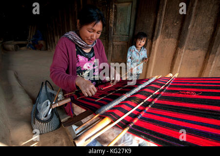 Donna tessitura tradizionale rosso naga scialle, giovane figlia accanto a lei, sulla veranda della casa di famiglia, Nagaland, India, Asia Foto Stock