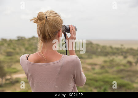 Turista femminile guardando attraverso il binocolo su african safari nel Serengeti National Park. tanzania, afrika. Foto Stock