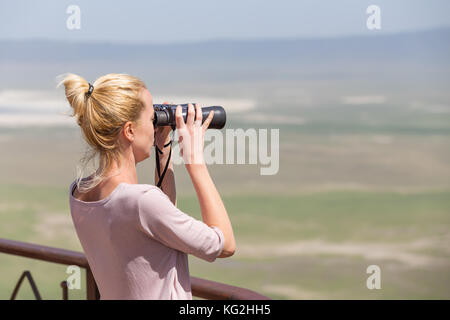 Turista femminile guardando attraverso il binocolo su safari africano nel cratere di Ngorongoro area consrvation, Tanzania, afrika. Foto Stock