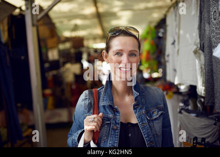 Felice attraente donna elegante shopping presso un mercato all'aperto in piedi guardando la telecamera con una vasta caloroso sorriso amichevole Foto Stock