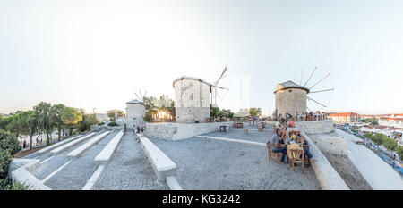 Vista panoramica ad alta risoluzione dei mulini a vento, dove la gente mangia e beve in un bar vicino in una giornata di sole nella città di Alacati, una destinazione popolare per i viaggi Foto Stock