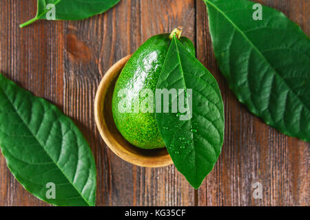 Un verde mature crudo intero frutto di Avocado consiste in una ciotola di legno, circondato da foglie su un tavolo marrone Foto Stock