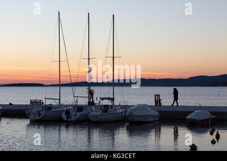 Un porto sul lago al tramonto, con varie barche e una donna a parlare al telefono mentre si cammina Foto Stock