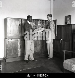 Degli anni Cinquanta, foto storiche che mostra un uomo britannico in tuta presentando un lungo riproduzione dischi in vinile,un Decca LP, ad un visitatore d'oltremare, Inghilterra, Regno Unito. Una cornice per una foto di una giovane regina Elisabetta è seduta su di un armadio. Foto Stock