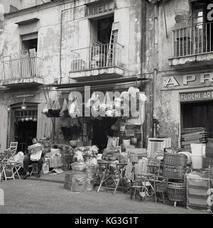 Degli anni Cinquanta, foto storiche di un Italiano stallholder leggendo il suo giornale al di fuori del suo negozio con il suo homewares e oggettistica traboccante sul marciapiede e in strada, Napoli, Italia, Foto Stock