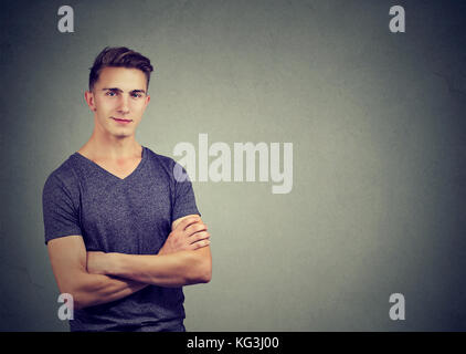 Casualmente bello fiducioso giovane uomo in t-shirt mantenendo le braccia incrociate mentre in piedi contro la parete sullo sfondo Foto Stock