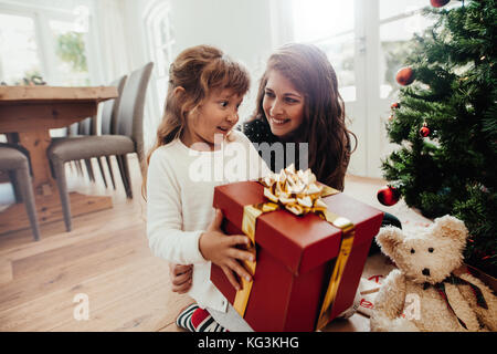 Bambina azienda regalo di Natale seduto con sua madre. La famiglia felice con doni seduta vicino ad albero di Natale a casa. Foto Stock