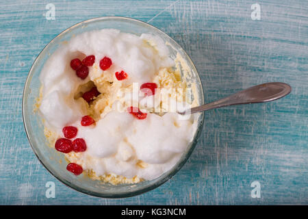 Impasto per cottage cheese casseruola, vista dall'alto. mantecato bianchi e bacche rosse di formaggio in una ciotola di vetro con un cucchiaio, un blu sullo sfondo di legno Foto Stock