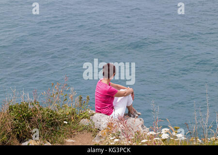 Spagna Cantabria - luglio 4, 2016: la donna sconosciuta sulla costa dell'oceano. la donna siede sulla costa alta e guarda lontano, una vista dal retro. sunny summ Foto Stock