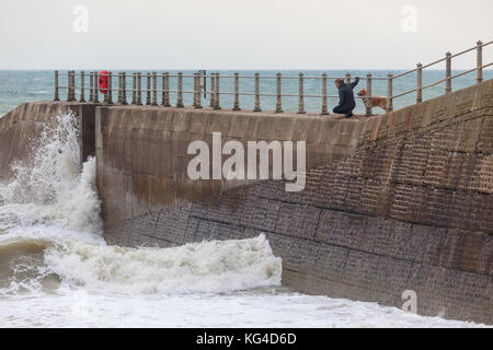 Hastings, East Sussex, Regno Unito. 4 Novembre, 2017. Condizioni di terreno accidentato fuori in mare in Hastings oggi con temperature di 14c e un 50% di probabilità di pioggia. Un uomo e il suo cane che guarda il mare dal mare muro di difesa come una grande onda crash. Photo credit: Paolo Lawrenson /Alamy Live News Foto Stock