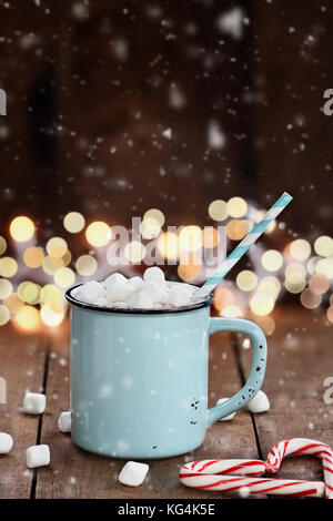 Smalto tazza di cioccolata calda con mini marshmallows e candy canne in forma di cuore contro un sfondo rustico con belle luci di Natale di Foto Stock