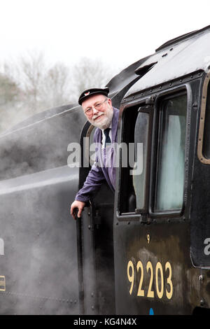 Un conducente si appoggia all'esterno della cabina con un getto di vapore treno che è temporaneamente fermata ad una stazione ferroviaria Foto Stock
