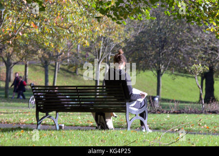 Giovane studentessa che studia a Kelvingrove Park giornata di sole seduta sulla panchina con le persone sullo sfondo viste da dietro Foto Stock