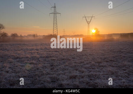 Linea ad alta tensione supporta alla mattina presto in campo il pupazzo di neve con erba vecchia fulgido di sunrise Foto Stock