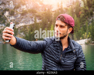 Giovane uomo bello tenendo selfie con lo smartphone al lago nella foresta. Foto Stock