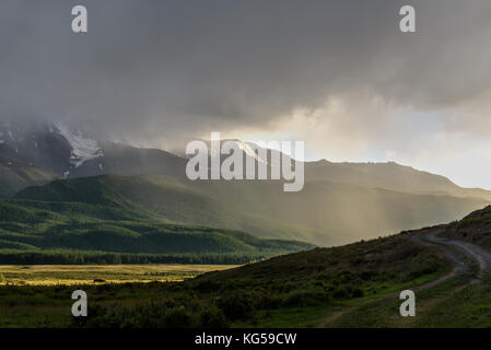 Vista panoramica con un lago e montagne coperte di neve, foresta, cielo nuvoloso, la nebbia e la pioggia al di sopra delle montagne con la luce solare Foto Stock