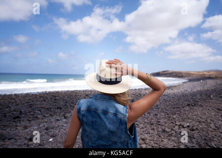 La donna torna con hat. che guardano sul mare. in piedi su una spiaggia di pietre. lanzarote isole canarie Foto Stock