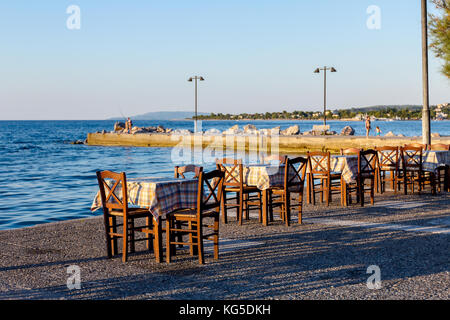 Sedie e tavoli in outdoor tipica taverna greca nella luce del tramonto con ombre vicino al molo. Foto Stock