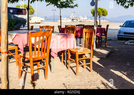 Sedie e tavoli in outdoor tipica taverna greca nella luce del sole di mattina con ombre vicino al molo. Foto Stock