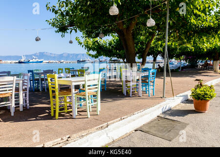 Sedie e tavoli in outdoor tipica taverna greca nella luce del sole di mattina con ombre vicino al molo. Foto Stock