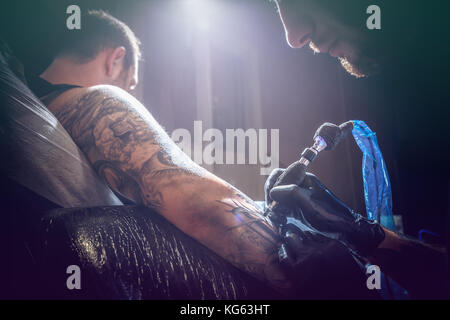 Ritratto di un uomo tattoo master che mostra il processo di creazione del tatuaggio in una mano d'uomo sotto la luce artificiale Foto Stock